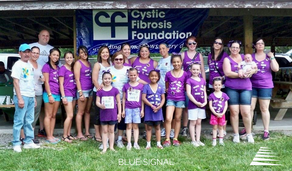 Group of people dressed in purple standing together in front of a sign reading Cystic Fibrosis Foundation, the recipient of this year's Blue Signal Gives Back donation