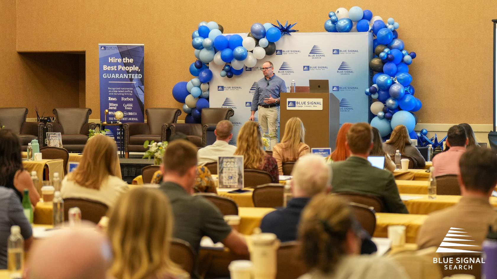 Keynote speaker addressing attendees at Blue Signal Summit 2024 with blue balloon decor and branded backdrop.